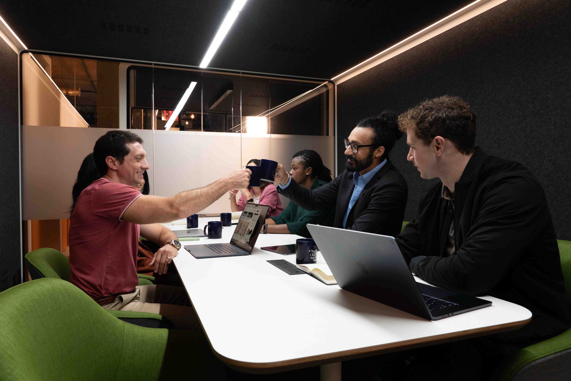 Two employees in a meeting booth with four other coworkers raise their coffee mugs in a friendly toast.