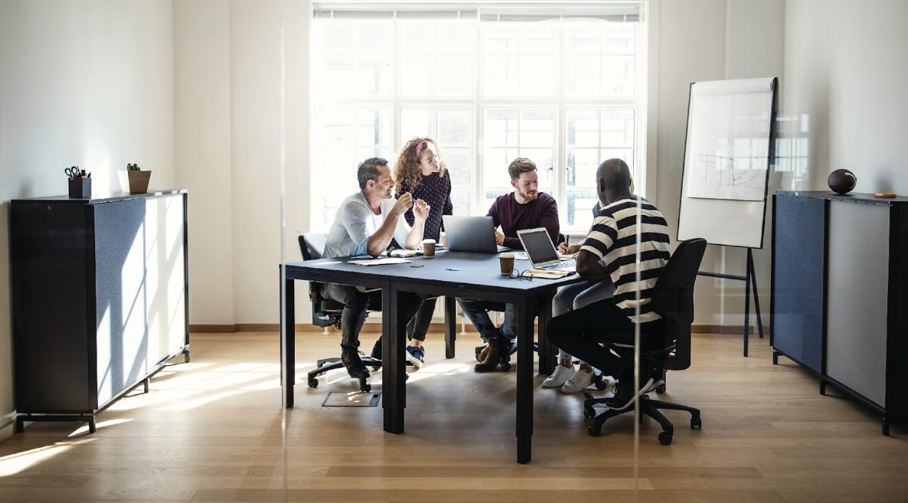  A group of colleagues collaborating in a huddle room, working on laptops and discussing ideas around a table.