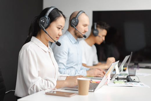 Three employees seated at a desk with their laptops. Each person is wearing a headset.