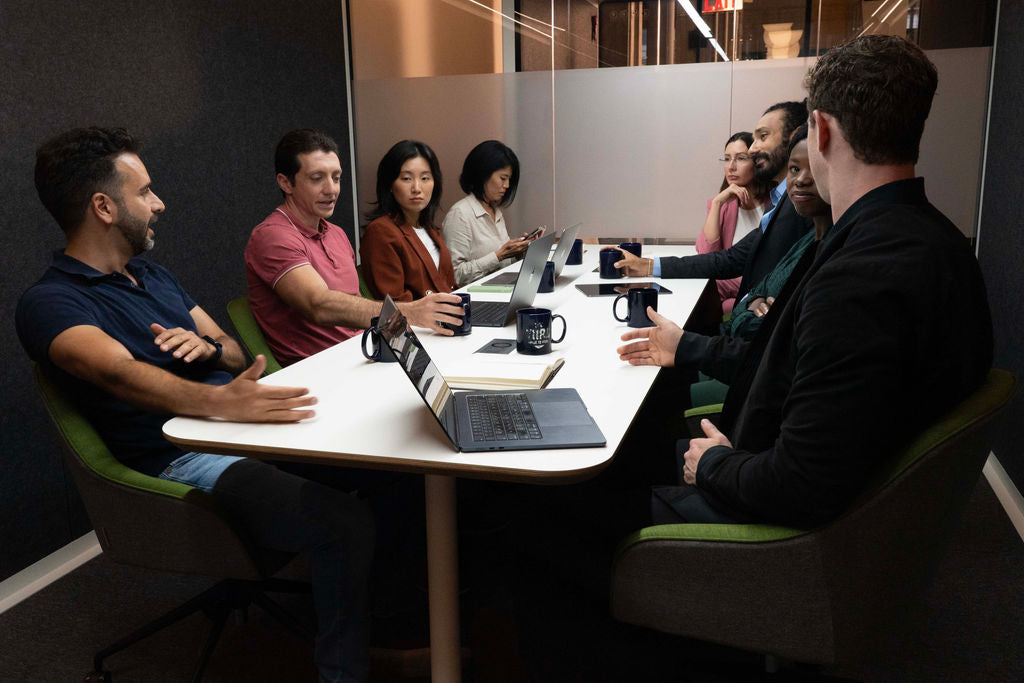 Coworkers seated at a desk with their coffee cups and laptops during a meeting