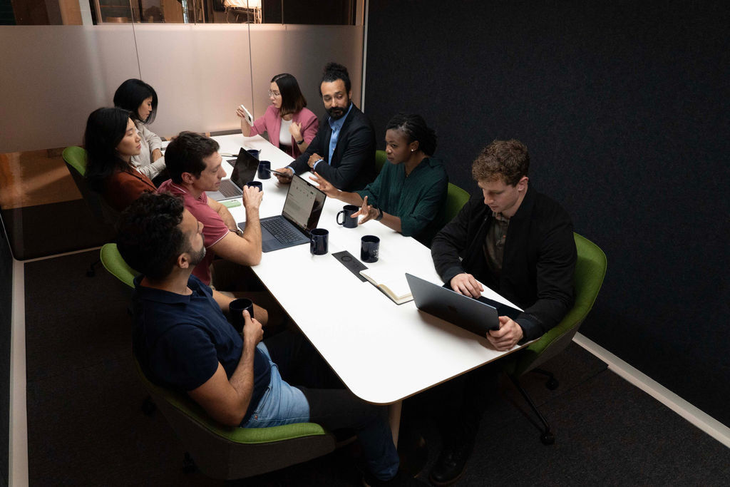 A team of coworkers seated at a conference table with their laptops and coffee mugs prepare for a meeting